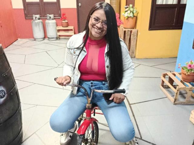 DahianaColeman, wearing glasses, a white jacket, red sheer top, and blue jeans, sits on a red bicycle, smiling, with long black hair, showing her tits.