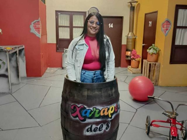DahianaColeman with long black hair, glasses, red top, blue jeans, white jacket, stuck in a wooden barrel labeled "Xarap deo's", smiling, in a colorful, quirky room.
