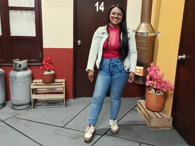 DahianaColeman has long black hair, wears glasses, a white jacket, red turtleneck, blue jeans, and white platform shoes. Her ass is small and she's smiling.