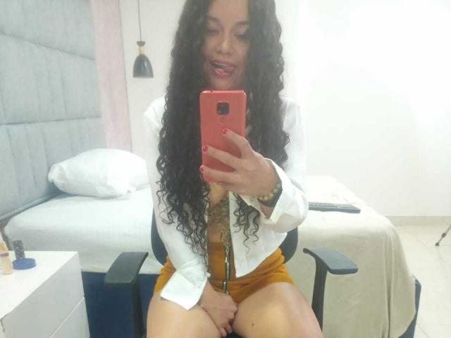 NatallyFlame with curly black hair, red lipstick, white shirt, and yellow shorts, sitting on a chair, covering her pussy with one hand, taking a selfie with a red phone.