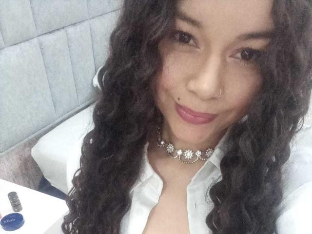 NatallyFlame with curly black hair, nose ring, and pink lipstick, wearing a white shirt and floral choker, smiling at the camera, her tits slightly visible under the shirt.