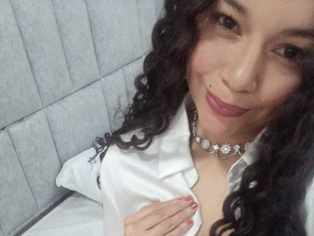 NatallyFlame with curly black hair, light brown skin, and a nose piercing, wearing a white shirt, floral necklace, and red lipstick, smiling at the camera, touching her chest.