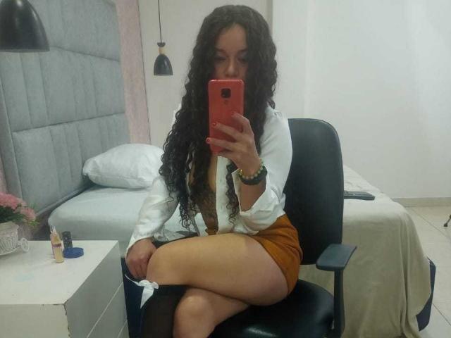 NatallyFlame with curly black hair, white blouse, brown skirt, and crossed legs, taking a mirror selfie with a red phone, showing her tits and ass in an office chair.