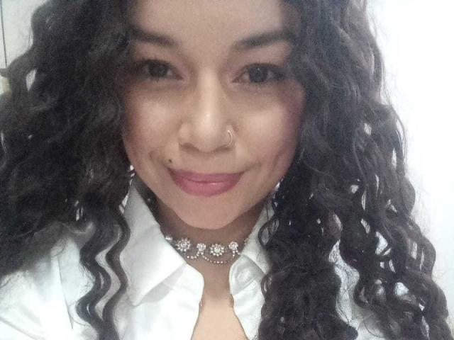 NatallyFlame with curly black hair, nose ring, mole under her right eye, pink lipstick, white shirt, and a sparkly choker necklace, smiling at the camera.
