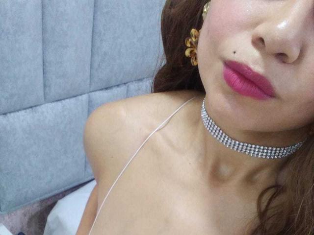 NatallyFlame with pink lips, brown hair, yellow flower earrings, diamond choker, and white strap, showing bare shoulders and a small mole on her face. Boobs not visible.