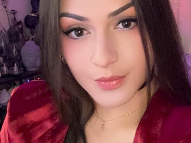 ElinaJhonson with dark brown hair, thick eyebrows, black eyeliner, pink eyeshadow, glossy pink lips, red velvet robe, gold necklace, and a nose ring, looking at the camera.