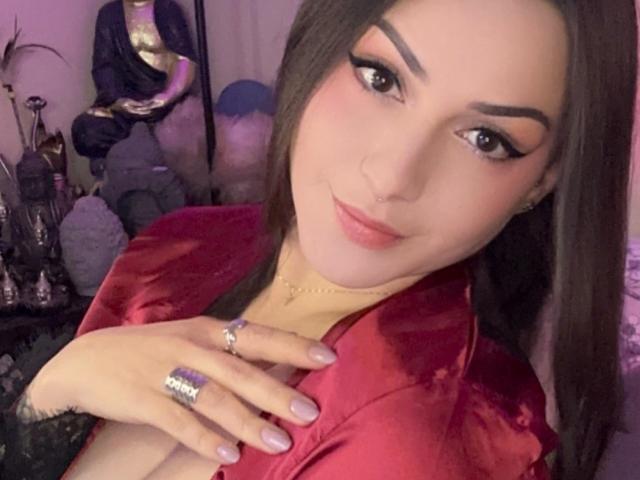 ElinaJhonson with dark hair, pink lips, and thick eyebrows, wearing a red velvet top, rings, and a nose ring, touching her chest with pink-nailed hands.