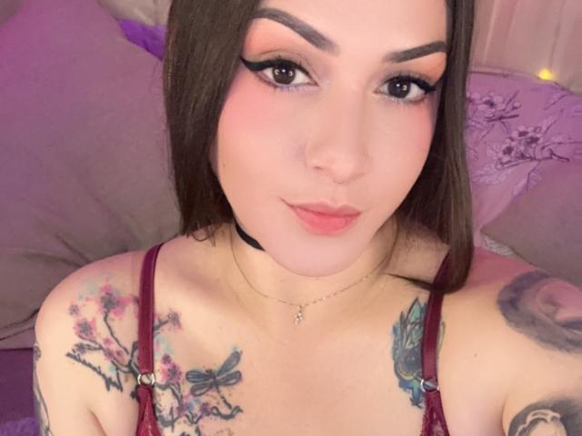 ElinaJhonson with tattoos on her tits and arms, wearing a red bra, has pink eyeshadow, dark eyebrows, and a black choker, smiling at the camera.