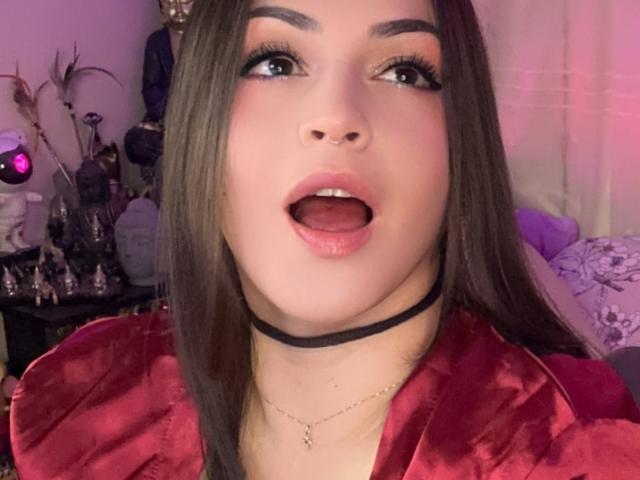 ElinaJhonson's face, mouth open, tongue slightly out, wearing a black choker, red satin top, long brown hair, eyeliner, and a nose ring. She has full, glossy pink lips.