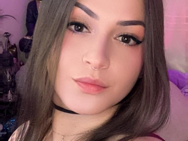 ElinaJhonson has brown eyes, thick eyebrows, long brown hair, a nose ring, and pink lipstick. She's wearing a black choker and a necklace. Her ass is not visible.
