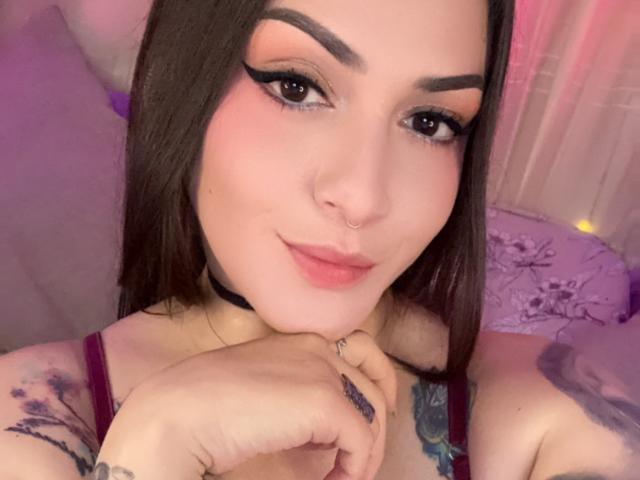 ElinaJhonson with pink makeup, black choker, nose ring, and tattoos, smiling at the camera, hands under chin, brown hair, dark eyebrows, wearing a purple top, soft pink lighting.