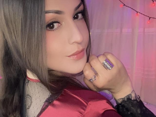 ElinaJhonson with long brown hair, pink lipstick, and brown eyes, wearing a red satin top with black lace, holding a ringed hand near her face, smiling slightly.