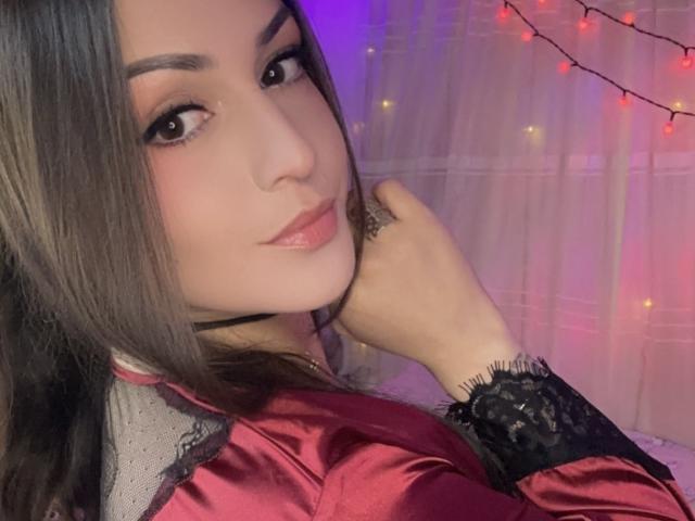 ElinaJhonson with long brown hair, pink lipstick, and a sexy gaze, wearing a red satin robe with black lace cuffs, her hand gently touching her chin.