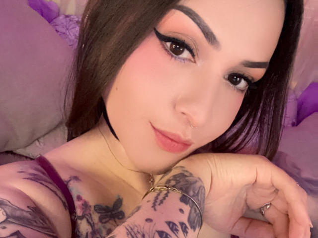 ElinaJhonson has tattoos on her arms, a nose ring, and dark eyeliner with pink eyeshadow. She's looking at the camera with a slight smile. Her tits are partially visible.