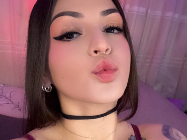 ElinaJhonson has pink lipstick, black eyeliner wings, a nose ring, and a black choker. She's puckering her lips and has long brown hair. Her tattoo is partially visible.
