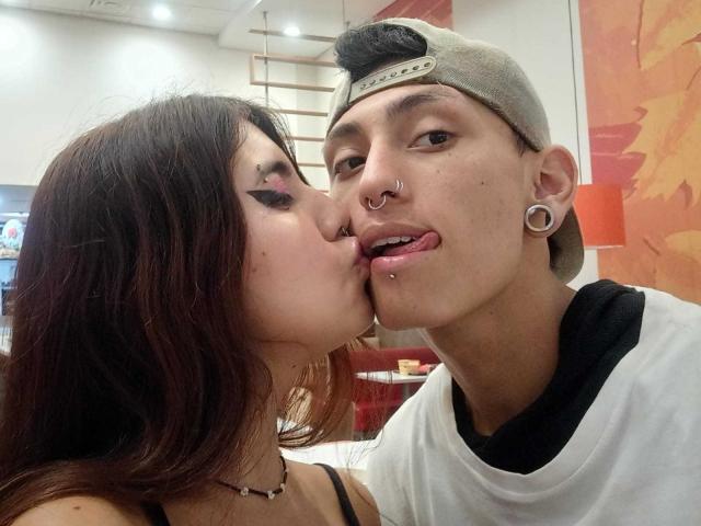 DainaAndMaik kissing, Daina with black eyeliner and nose ring, Maik with white tee, backward cap, nose ring, and ear gauges, tongue out, in a brightly lit room.