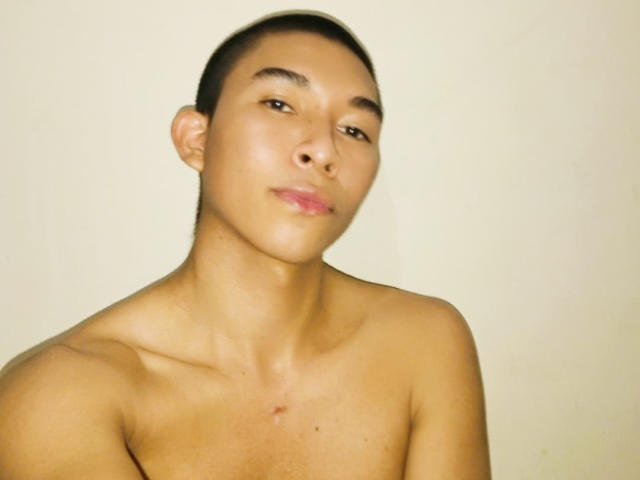 NohaKerry, shirtless with a smooth, tan chest, short brown hair, and a small scar below the collarbone, looking slightly to the side with a relaxed, slightly smirking face.
