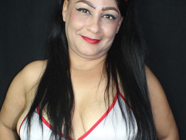 A CandelaFoxy tem cabelos pretos longos, batom vermelho e está sorrindo, com os seios visíveis em uma blusa branca com detalhes vermelhos, mostrando um pouco de decote contra um fundo preto..
