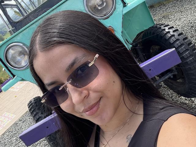 HikariChloe with long black hair, wearing dark sunglasses, smiling at the camera, has small tattoos on her chest, standing in front of a green, off-road vehicle with big tires.