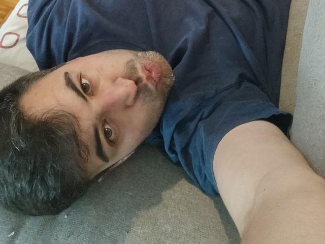 Vio69x is lying on a grey couch, head tilted back, dark eyebrows, short brown hair, and a slight beard, wearing a blue shirt, with a neutral expression.
