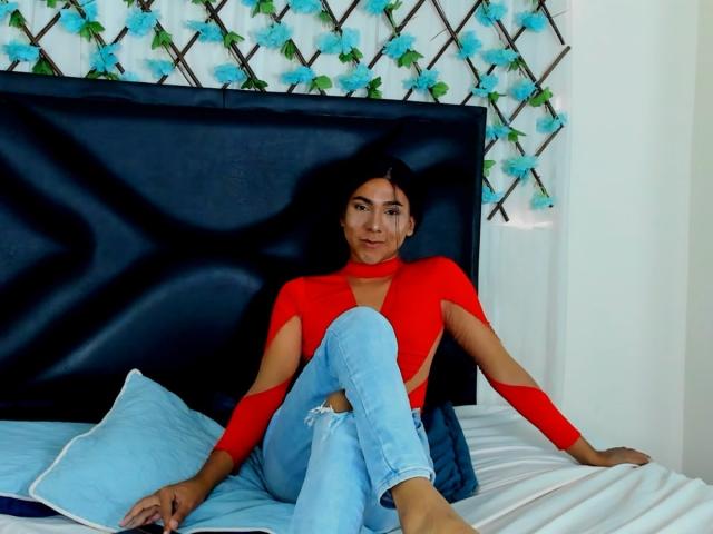 ZoeHotMoretti met kleine borsten, gekleed in een rode top met lange mouwen en gescheurde blauwe jeans, zittend op een bed met een zwart hoofdeinde, lachend naar de camera..