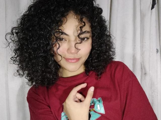 SammyHills69 has curly black hair, light brown skin, and a small nose ring. She's wearing a red shirt, pointing at her chest with a slight smile.