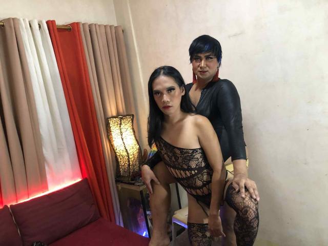 TwoBigCockShowTS with black lace outfit and medium tits, bent over, being held from behind by TwoBigCockShowTS in black shirt, in dimly lit room with red and beige curtains.