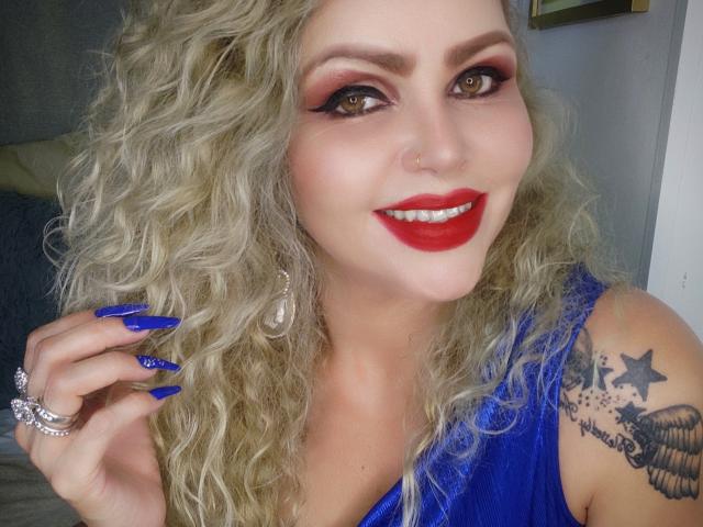 AnnaRichie with curly blonde hair, red lipstick, blue nails, blue dress, star tattoo, and ring, smiling at camera. She's got a nose ring and eye makeup.