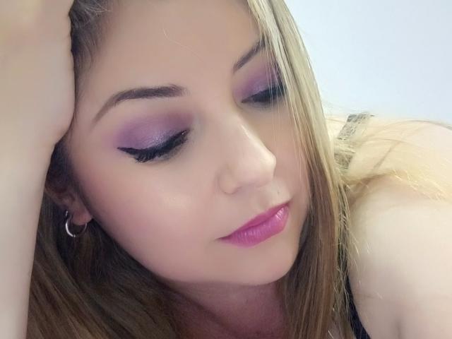 NatalyHami has purple eyeshadow, long blonde hair, pink lipstick, and a silver hoop earring. She's resting her head with closed eyes, looking sexy and relaxed.