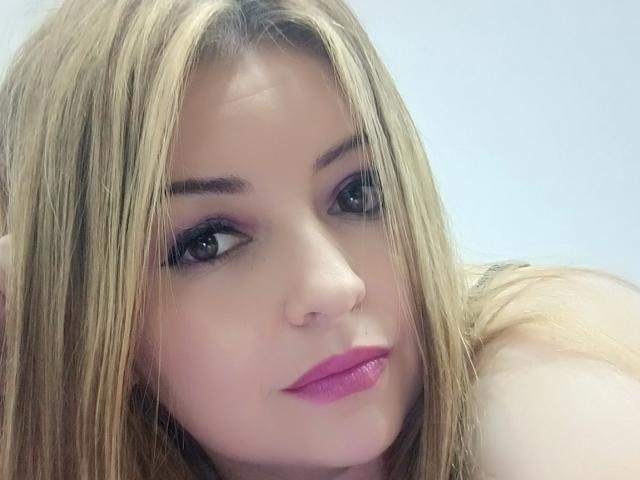 NatalyHami's face, blonde hair, purple eyeliner, black eyeshadow, pink lipstick, looking at camera, slight smile, fair skin, soft light, slightly messy hair, upper body visible.