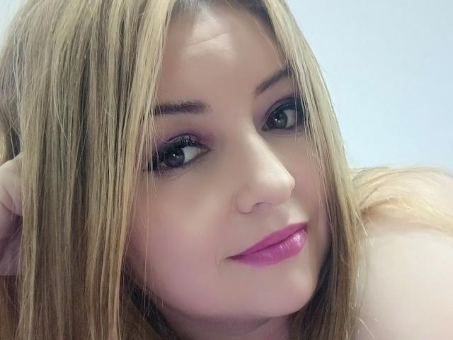 NatalyHami has blonde hair, purple eyeliner, and pink lipstick. Her face is slightly angled with a subtle smile. Her hand rests behind her head.
