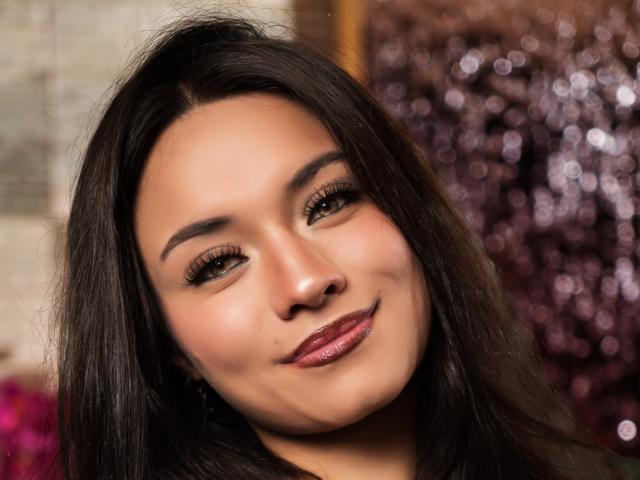 LaylaRousee's face with dark brown hair, brown eyes, and pink lipstick, smiling softly. She has a small mole on her left cheek and is wearing dark eyeliner.