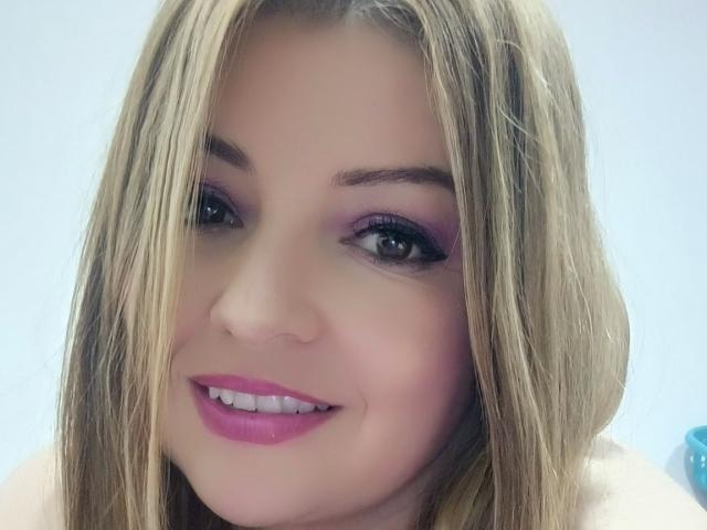 Het gezicht van NatalyHami, blond haar, roze lippenstift, paars oogschaduw, lachend, lichte huid, blauwe ogen, lichtjes open mond, zichtbare tanden, subtiele make-up, gecentreerd in de afbeelding..