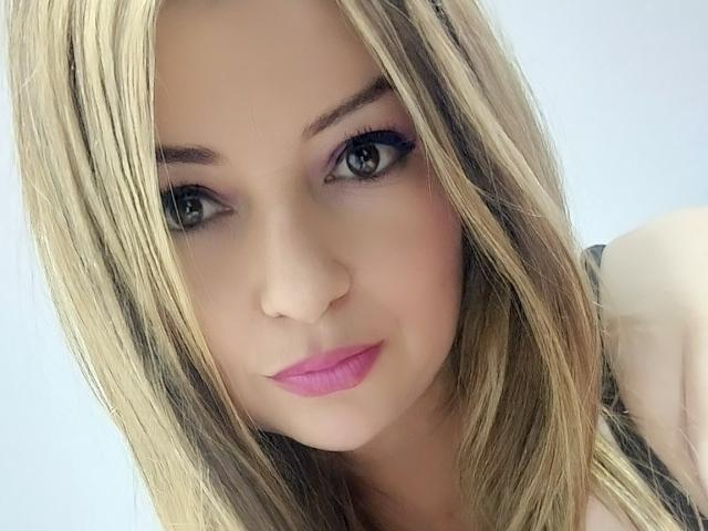 NatalyHami hat blondes Haar, pinken Lippenstift und grüne Augen mit schwarzem Eyeliner. Sie schaut mit einem leichten Lächeln in die Kamera und zeigt ihr süßes Gesicht und ihre Brüste..
