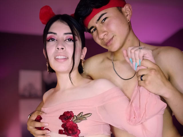 NoahAndRose with red pom-poms in hair, red headband, holding pink off-shoulder top with red roses, standing behind NoahAndRose with blue nails, smiling. NoahAndRose is shirtless.