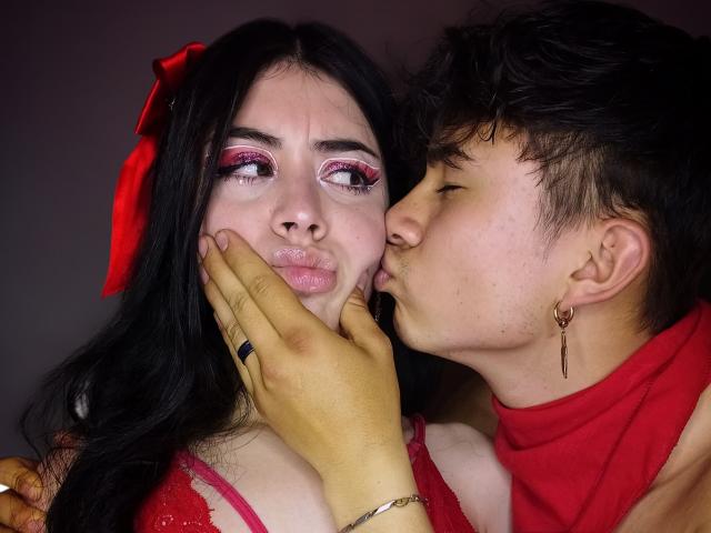 NoahAndRose with pink eyeshadow and red bow, being kissed on the cheek by NoahAndRose with short black hair, red shirt, and gold earring, holding her face gently.