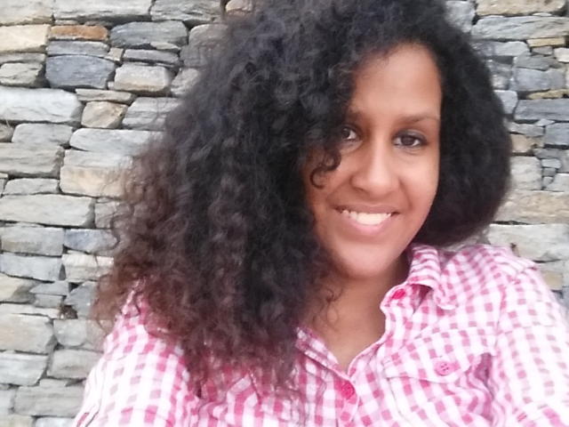 ShirliHot with curly black hair, brown eyes, and a smile, wearing a pink checkered shirt, has a cute face and small tits. Stone wall in background.