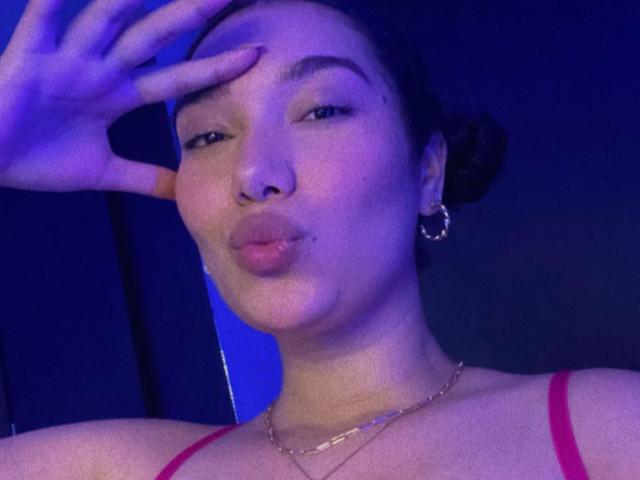 Ashlley69 with pouty lips, dark hair in bun, wearing red strap, gold necklace, hoop earrings, blue-tinted light, hand touching forehead, slightly squinted eyes, subtle makeup, and a small mole.