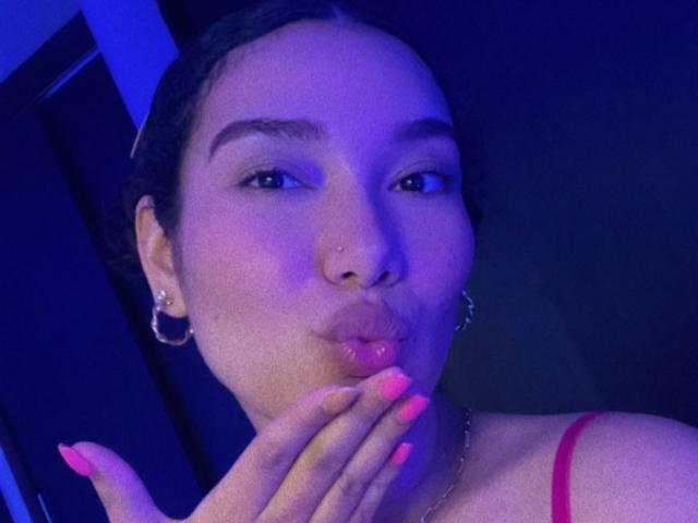 Ashlley69, con uñas rosas, labios carnosos y aros grandes, besando sus dedos, vistiendo una blusa roja, bajo una iluminación azul, mostrando un rostro lindo y sexy..