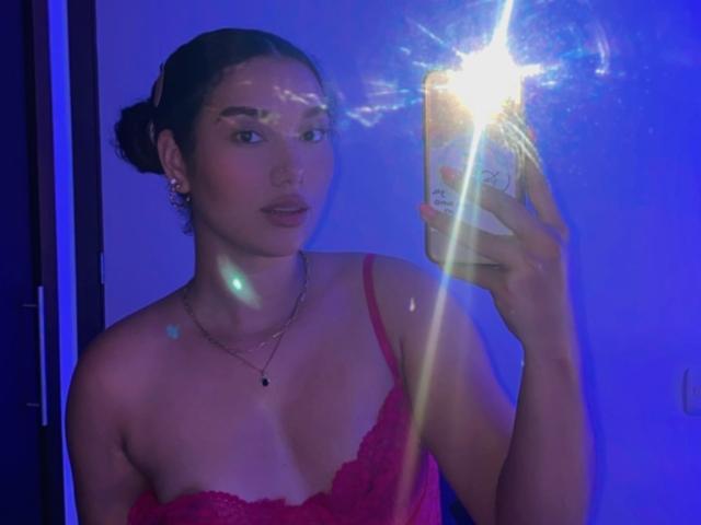 Ashlley69, com cabelo escuro preso em um coque, usando um sutiã de renda vermelho, segurando um celular com um flash forte, mostrando seus seios e rosto, em um quarto com iluminação azul..