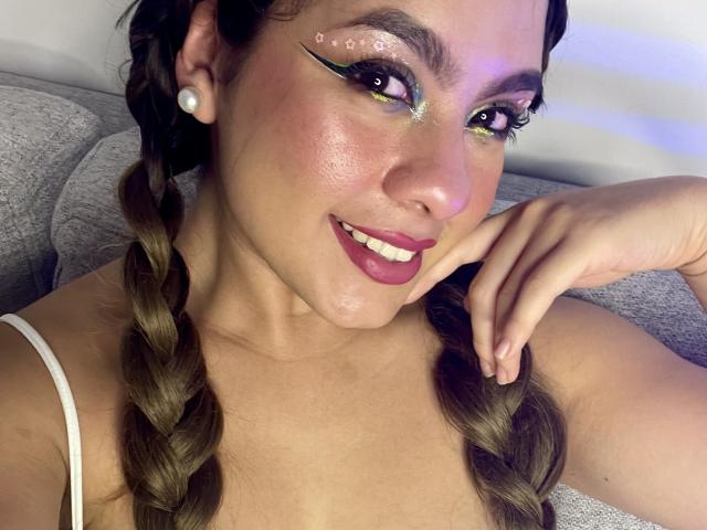 AllysonSexxy, met gevlochten bruin haar, draagt witte oorbellen, heeft groene oogschaduw, zwarte eyeliner, roze lippenstift en raakt met haar rechterhand haar kin aan, terwijl ze glimlacht..