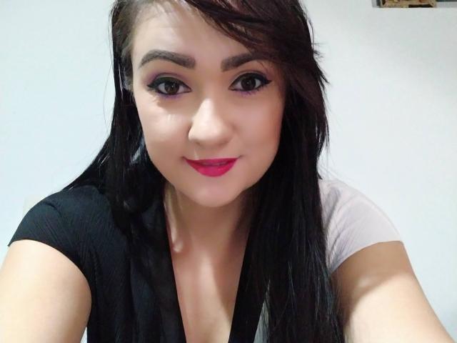 Katnnis with long black hair, purple eyeliner, red lipstick, and a black/white top, smiling at the camera, showing her face and upper chest.