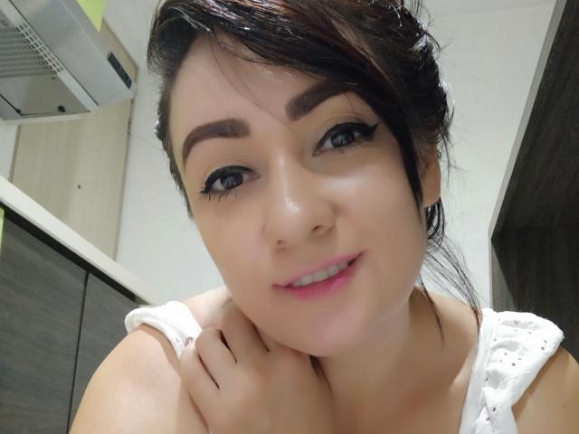 Katnnis with dark brown hair, black eyeliner, pink lipstick, and white sleeveless top, smiling slightly, hand near chin, in a kitchen. Her tits are not visible.
