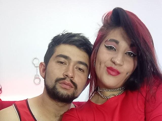 BonniClark with red hair, makeup, nose ring, and lip studs, next to a bearded man in a red shirt. Her tits are small, his dick is not visible.