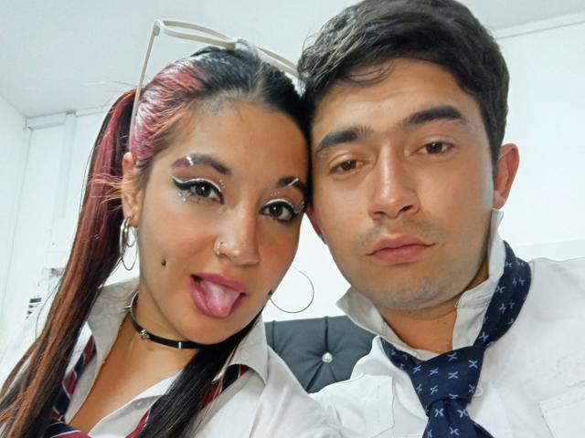 BonniClark with red ponytail and black choker, sticking out tongue, wearing white shirt, black necklace. Her man, brown hair, white shirt, blue tie, pouting. Both in selfie.