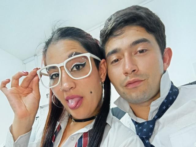 BonniClark with large white glasses, pink lipstick, and a nose piercing, sticking out her tongue. Next to her is a man in a white shirt and blue tie.