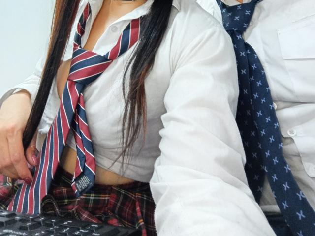 BonniClark with long black hair, white shirt, red-striped tie, small tits, plaid skirt, sitting next to someone in a white shirt and blue star-patterned tie.