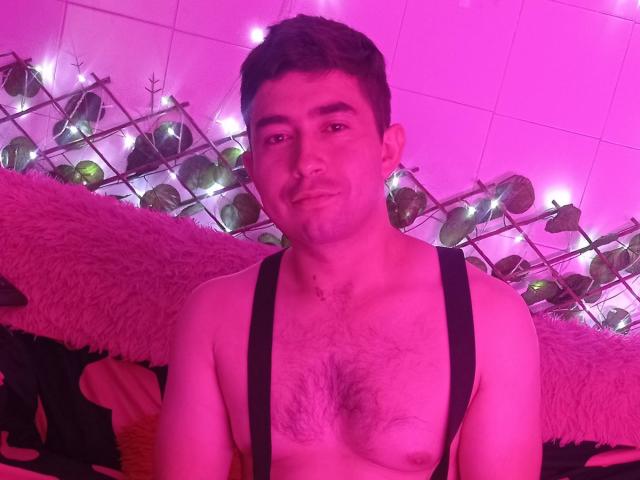 BonniClark, shirtless with black suspenders, has a hairy chest and short dark hair, under pink lighting with string lights and green plants in the background.