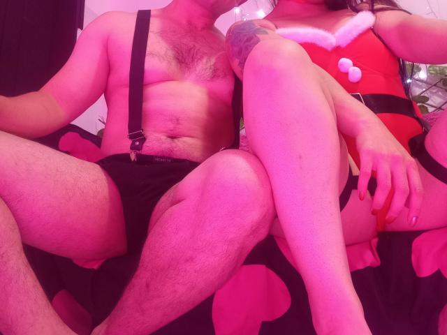 BonniClark with hairy chest and black shorts, sitting next to BonniClark in red Santa outfit, showing bare legs and tattoo on arm, under pink lighting.