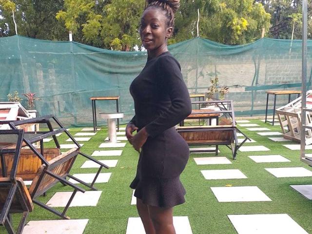 CaramelAnna69 with dark skin, curly hair in a bun, wearing a black long-sleeve dress and ruffled skirt, smiling, showing her ass, standing on green grass.
